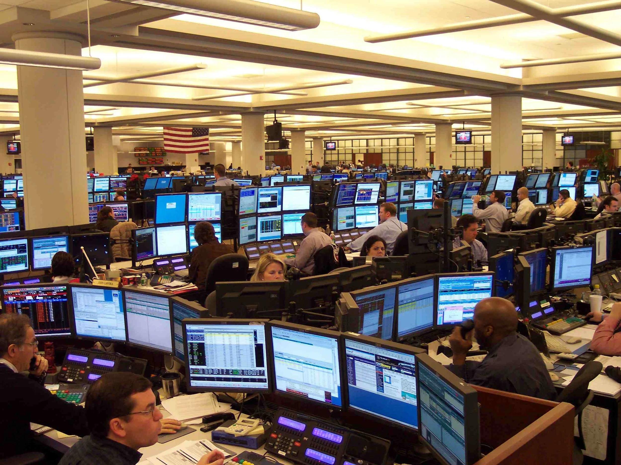 A modern trading floor with rows of trading desks, computer monitors and a central market-display screen.